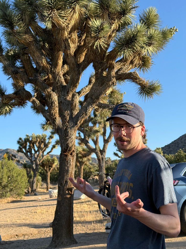 Riley Strader, founder of Dead Letter, mid-shrug in the desert in front of a Joshua tree